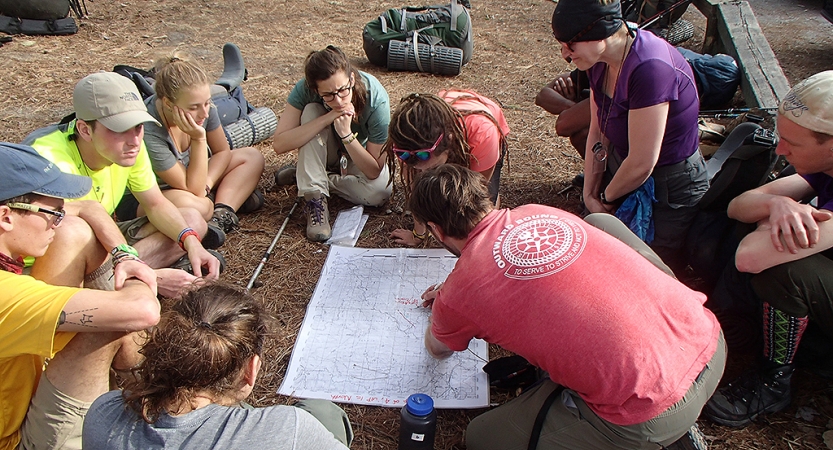 A map lays on the ground and people sit around it. One person, who appears to be an instructor, points at the map.
